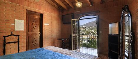 Una camera da letto con un letto, una porta in legno, un tavolino, uno specchio e vista su un paese attraverso una porta aperta.
