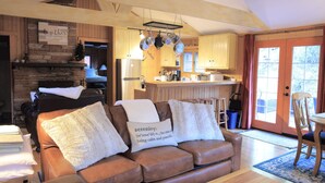 Een gezellige woonkamer met een bruine leren bank, een houten plafond en een keuken met houten kastjes en een roestvrijstalen koelkast.
