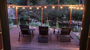 Een houten terras met drie rieten stoelen en een kleine tafel, met uitzicht op een natuurlandschap met een beekje en bomen.
