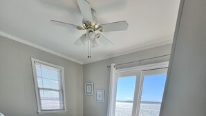 Un dormitorio con cama, ventilador de techo, ventana con persianas y una pequeña mesa con lámpara, con vista a la playa.