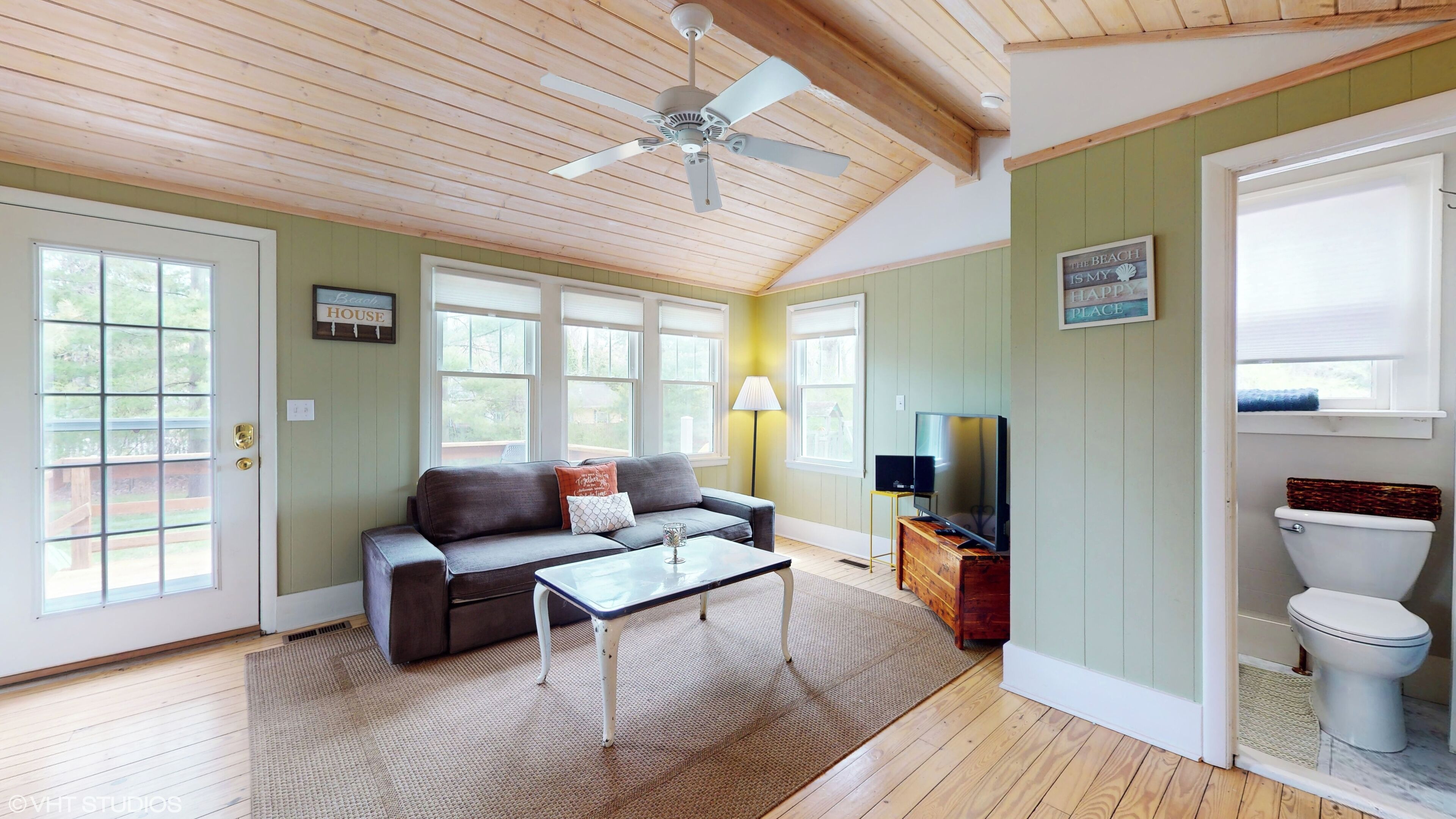 A spacious living room with a ceiling fan, a sofa, a coffee table, and a television. There are large windows and a door leading to the outside.