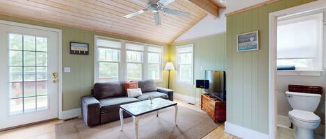 A spacious living room with a ceiling fan, a sofa, a coffee table, and a television. There are large windows and a door leading to the outside.