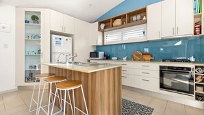 A modern kitchen with a central island, bar stools, a microwave, a stove, and a built-in oven. - White Sands Deck House by Experience Jervis Bay (Huskisson)