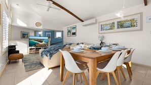 A modern dining area with a wooden table set for four, white chairs, and a view of the living room in the background. - White Sands Deck House by Experience Jervis Bay (Huskisson)