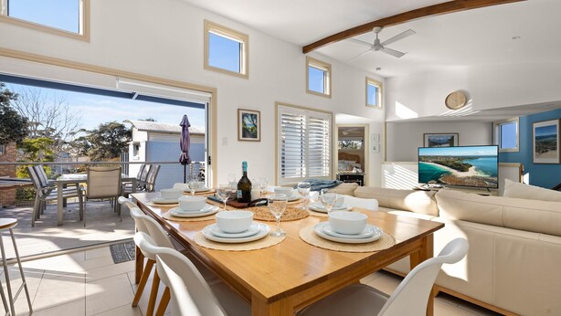 A spacious dining area with a wooden table set for a meal, a sofa, and a television displaying a scenic view. - White Sands Deck House by Experience Jervis Bay (Huskisson)
