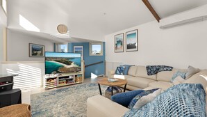 A living room with a large flat-screen TV, a sofa, a coffee table, and a bookshelf. - White Sands Deck House by Experience Jervis Bay (Huskisson)