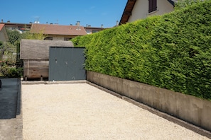 A gravel driveway leading to a wooden gate, a neatly trimmed hedge, and a residential building with a tiled roof in the background.