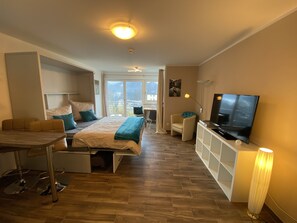 A modern bedroom with a bed, a desk, a chair, a TV, and a large window.