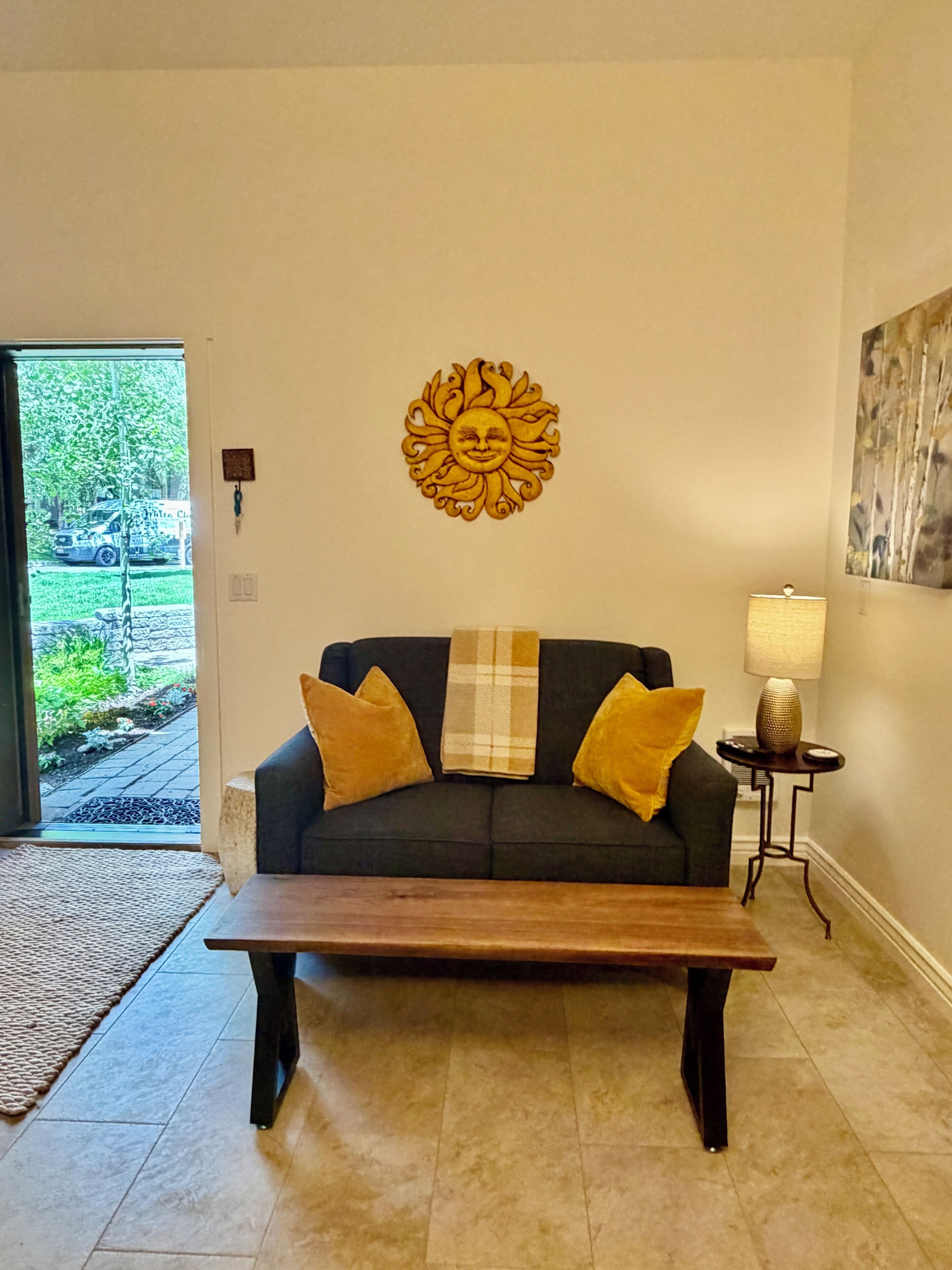 Un salon avec un canapé de couleur foncée, deux coussins jaunes, un plaid, une table basse en bois, une décoration murale représentant un soleil, un lampadaire et une porte donnant sur l’extérieur.