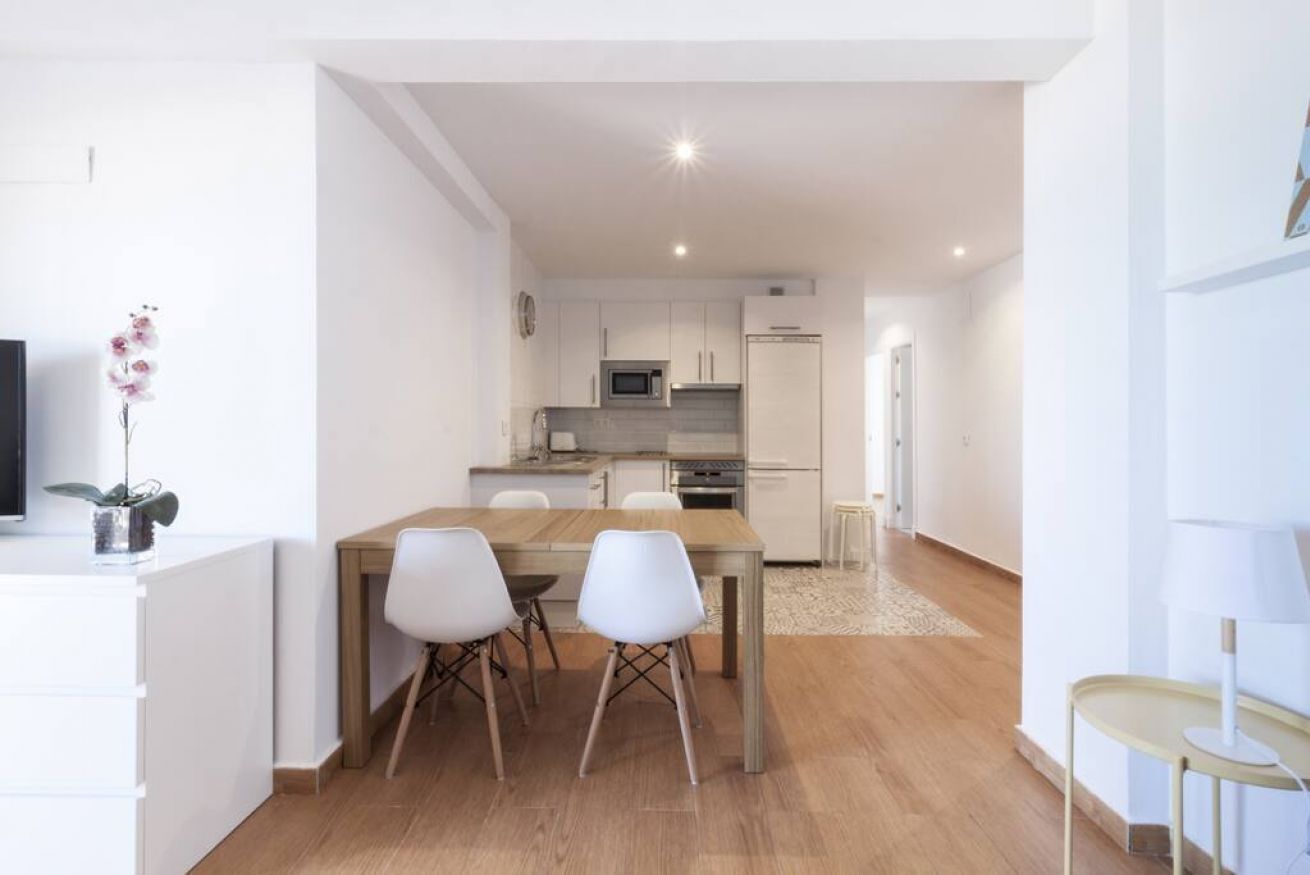 A modern kitchen with white cabinets, a microwave, and a refrigerator. There is a dining area with a wooden table and white chairs.