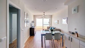 A modern living room with a dining area, a sofa, a ceiling fan, and a view of the outdoors. - Apartamento Playa Victoria Cadiz (Cádiz)