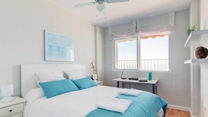 A bedroom with a bed, a desk, a chair, a ceiling fan, a window with curtains, and a wall-mounted shelf. - Apartamento Playa Victoria Cadiz (Cádiz)