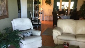 A living room with a white armchair, a beige sofa, a glass coffee table, and a large green plant. - Close To Anna Maria Island, Beaches. On IMG Golf Course. Short Drive To Sarasota (Bradenton)