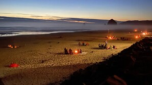 Una spiaggia al crepuscolo con persone riunite attorno a dei fuochi, tende e una grande formazione rocciosa in lontananza.