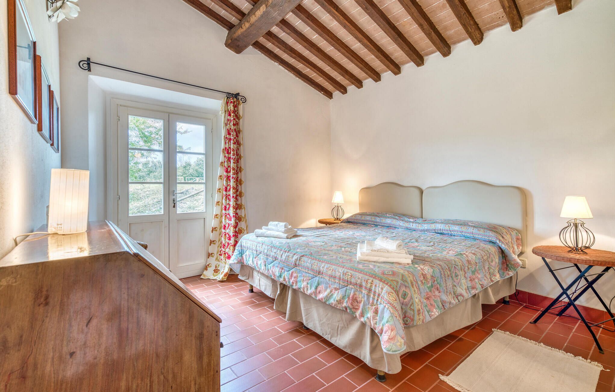 Una camera da letto con un letto, due comodini con lampade, una finestra con tende, una scrivania in legno e un pavimento piastrellato.