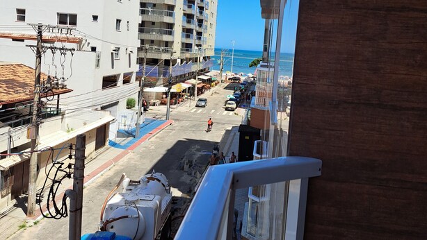 A view from a balcony overlooking a street with buildings, a clear blue sky, and a distant view of the sea. - Apartment 200m beach (with view), rest and comfort, close to everything. (Praia do Morro)
