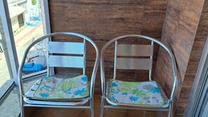 Two metal chairs with floral cushions on a tiled balcony with a wooden wall and a window overlooking a residential area. - Apartment 200m beach (with view), rest and comfort, close to everything. (Praia do Morro)