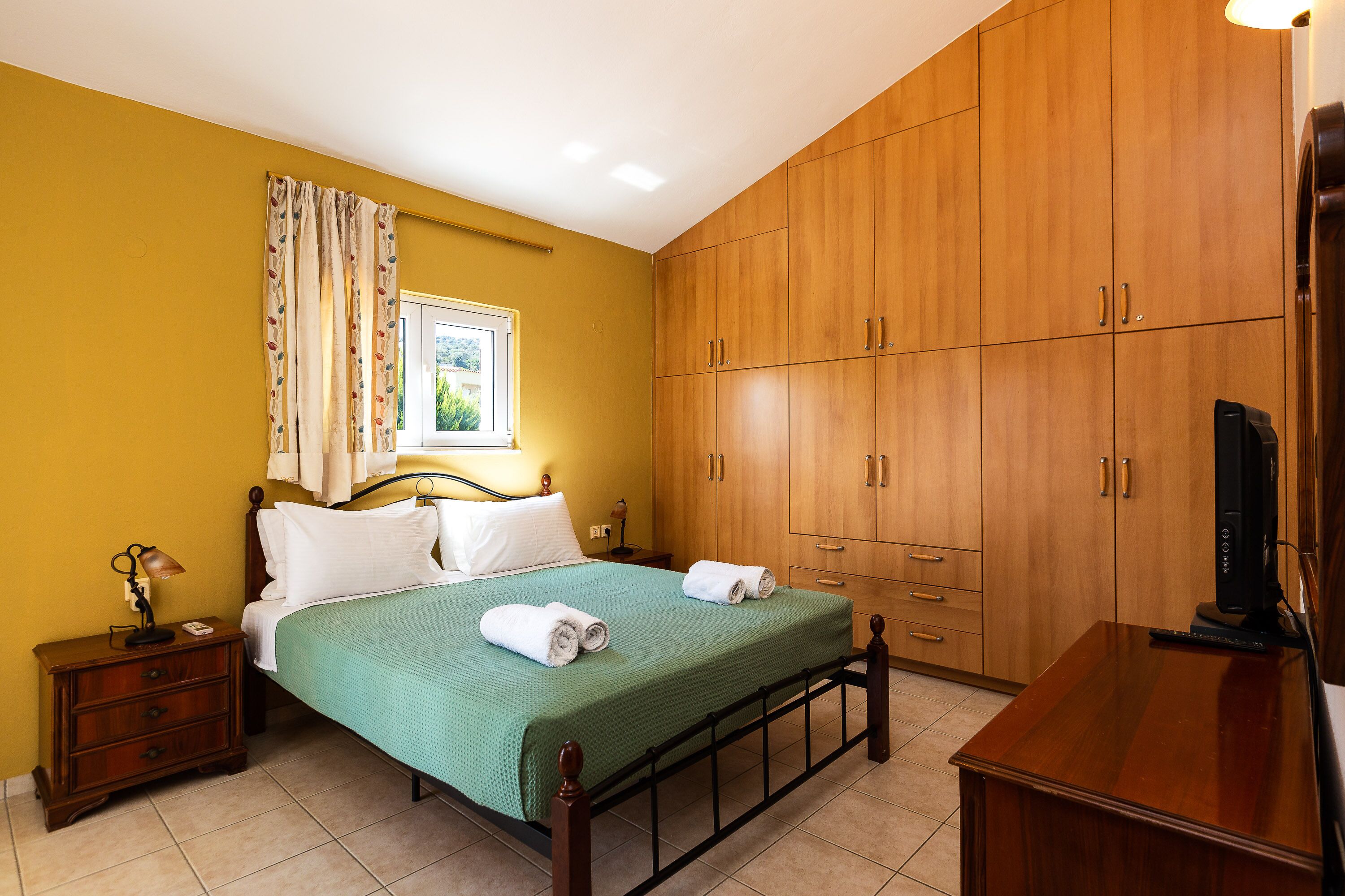 A bedroom with a bed, wooden wardrobe, and a desk with a television.