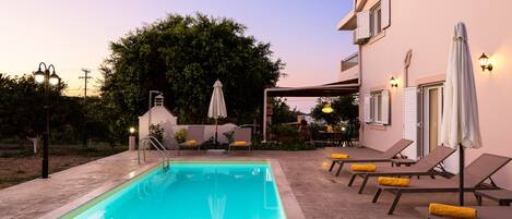 A swimming pool with lounge chairs and umbrellas, surrounded by a building with outdoor seating and a tree.