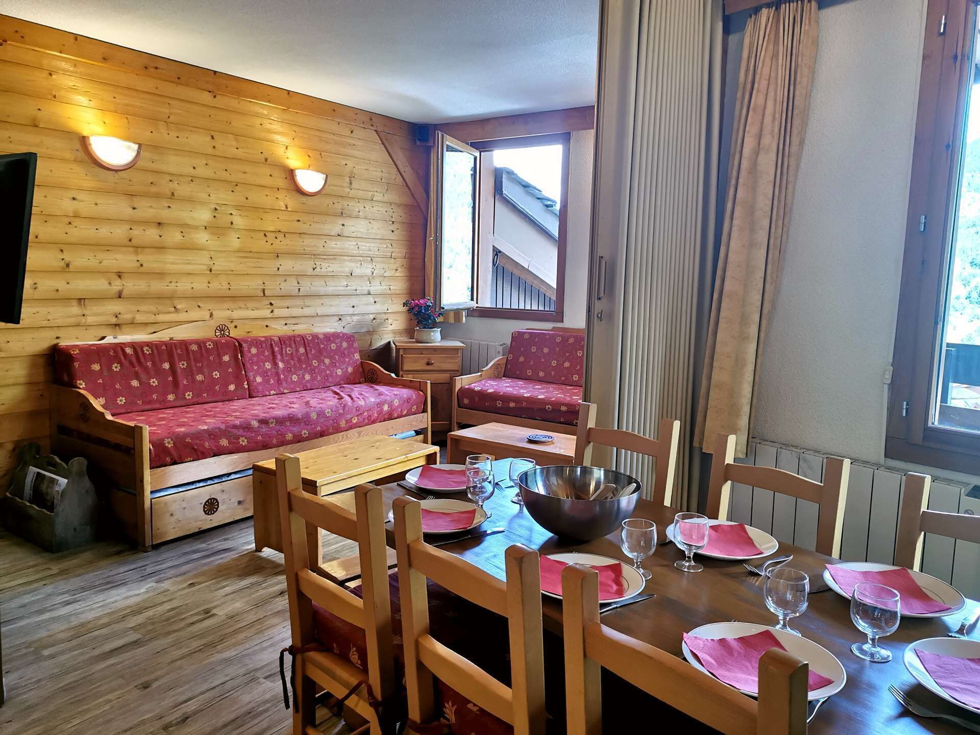 Interior acogedor de una cabaña con pared de madera, un sofá, una mesa de comedor para cuatro personas y una ventana con cortinas.
