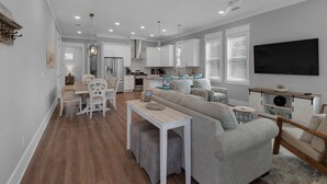 A modern kitchen with white cabinets, a dining area with a table and chairs, a gray sofa, and a flat-screen TV mounted on the wall. - Grayton Beach Vacation House- park your car, hop on a bike, explore! (Santa Rosa Beach)