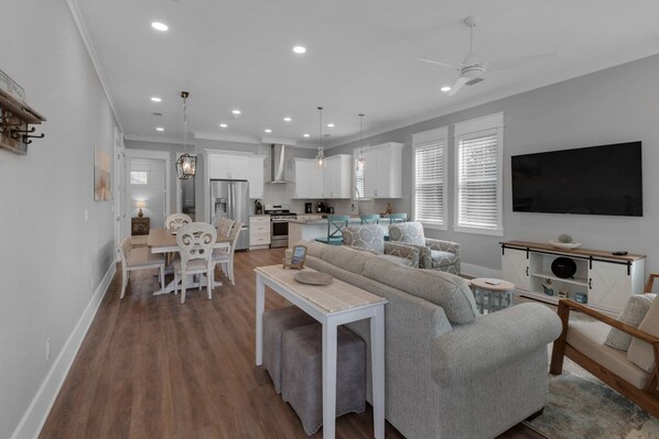 A modern kitchen with white cabinets, a dining area with a table and chairs, a gray sofa, and a flat-screen TV mounted on the wall.