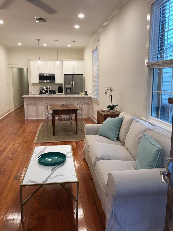 A modern living room with a wooden floor, a white sofa with teal cushions, a glass coffee table, and a kitchen area with white cabinets and stainless steel appliances.