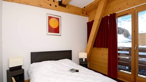 A bedroom with a large bed, wooden ceiling beams, a nightstand with a lamp, a framed picture on the wall, and a view of a snowy landscape through a sliding door.