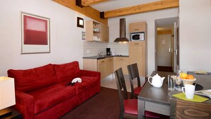 A compact living space with a red sofa, a dining table with chairs, a kitchen area with a microwave, and a framed picture on the wall.