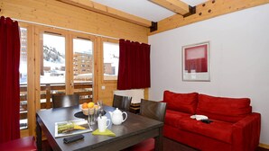 A cozy room with a wooden ceiling, a red sofa, a dining table with chairs, and a window with red curtains.