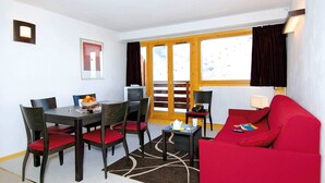 A room with a dining table, four chairs, a red sofa, a small side table, a lamp, and a window with curtains.