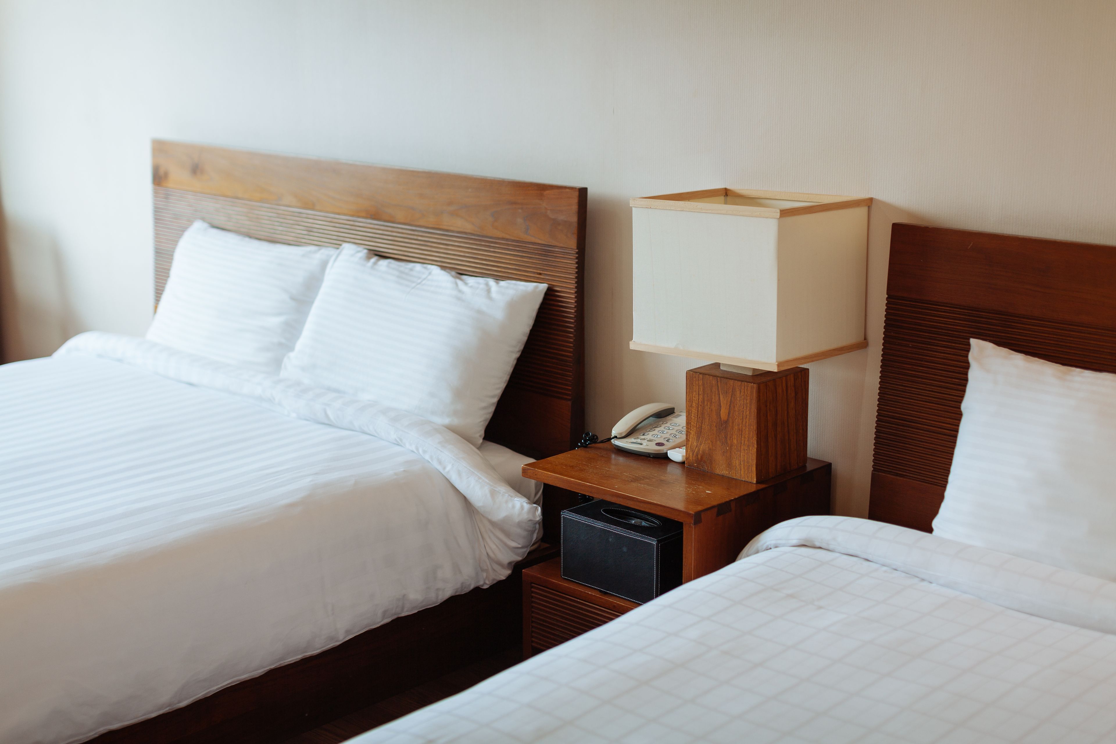 A hotel room with two beds, each with white bedding and wooden headboards. There is a bedside table with a lamp and a telephone on one side.