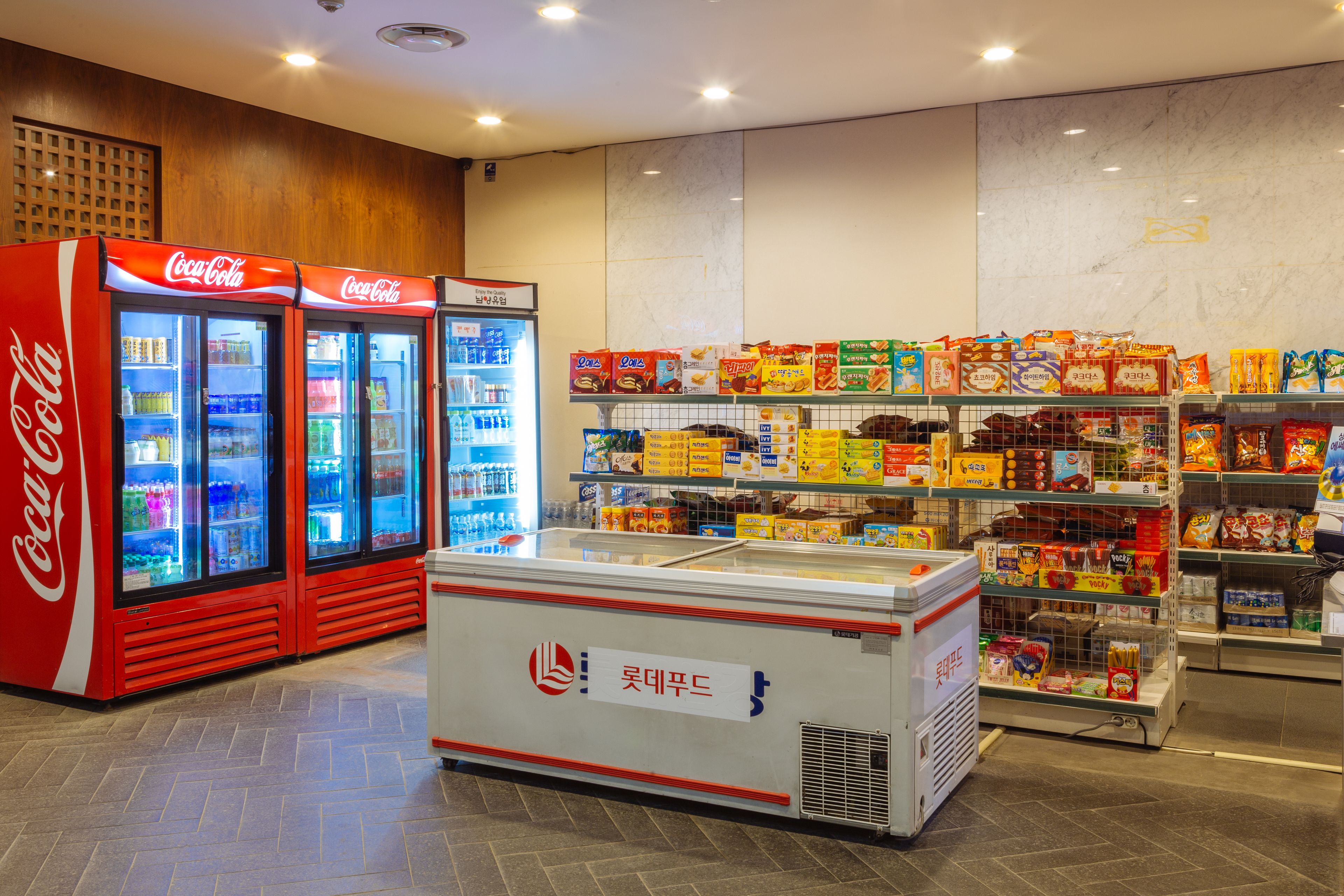 Zona de exhibición refrigerada con enfriadores de Coca-Cola y un congelador, rodeada de estantes llenos de diversos productos envasados.