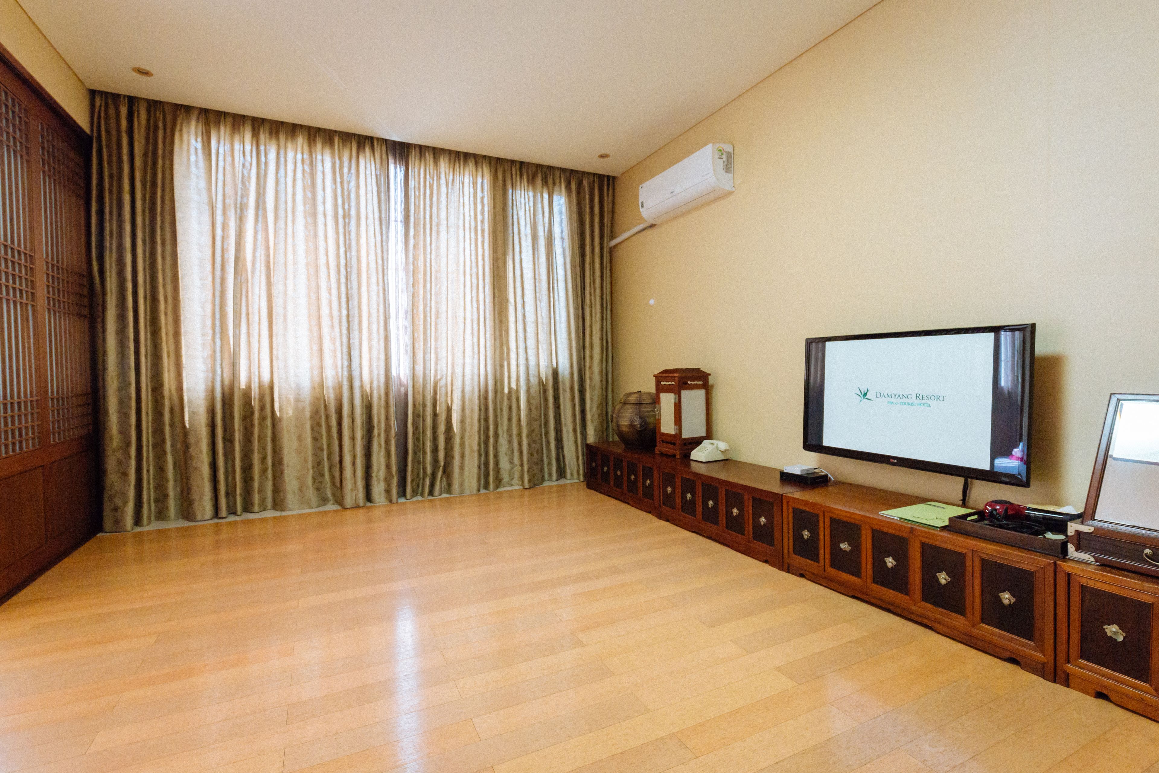 Una habitación amplia con piso de madera, un ventanal con cortinas, un televisor de pantalla plana sobre un mueble de madera y un equipo de aire acondicionado en la pared.