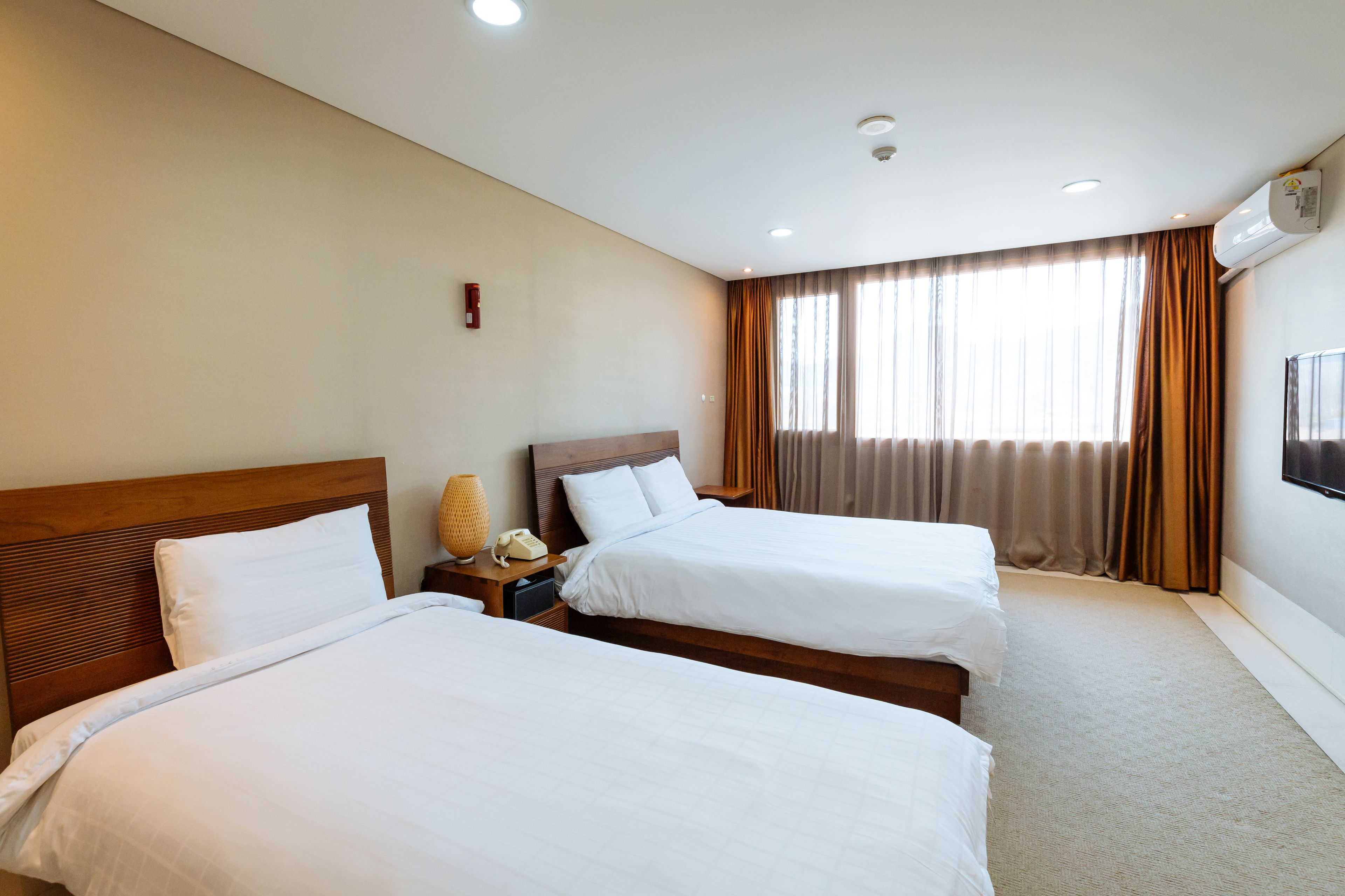 A hotel room with two beds, a bedside table with a lamp, a wall-mounted TV, and a window with curtains.