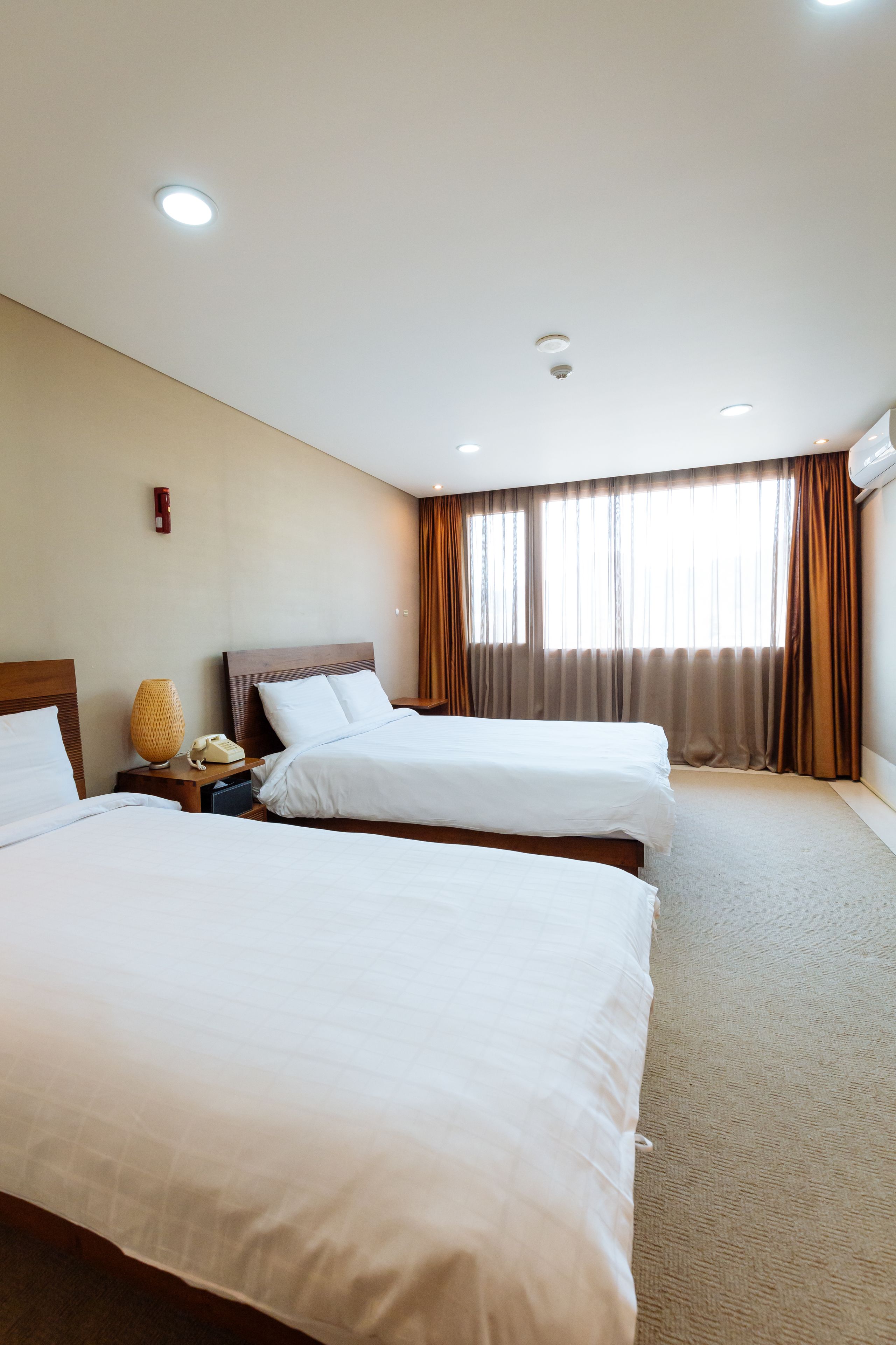 A hotel room with two beds, a bedside table with a lamp and a telephone, a window with curtains, and a ceiling light.