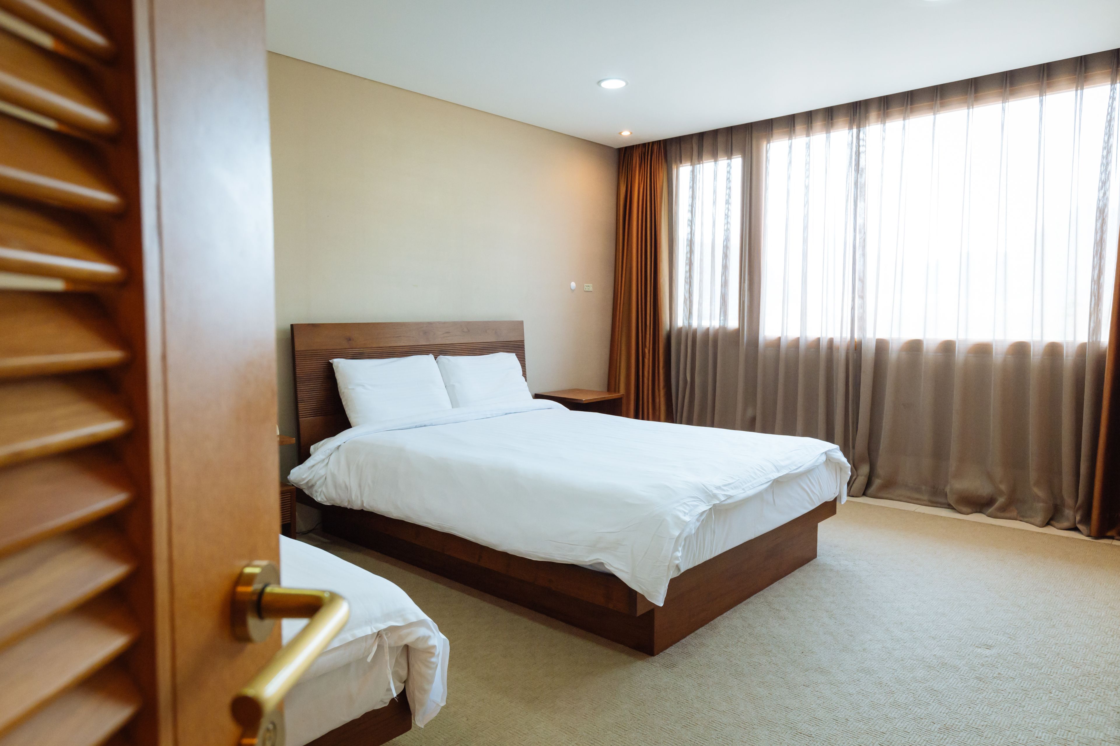 Een net opgemaakt bed in een hotelkamer met een houten hoofdeinde, een nachtkastje en een raam met gordijnen.