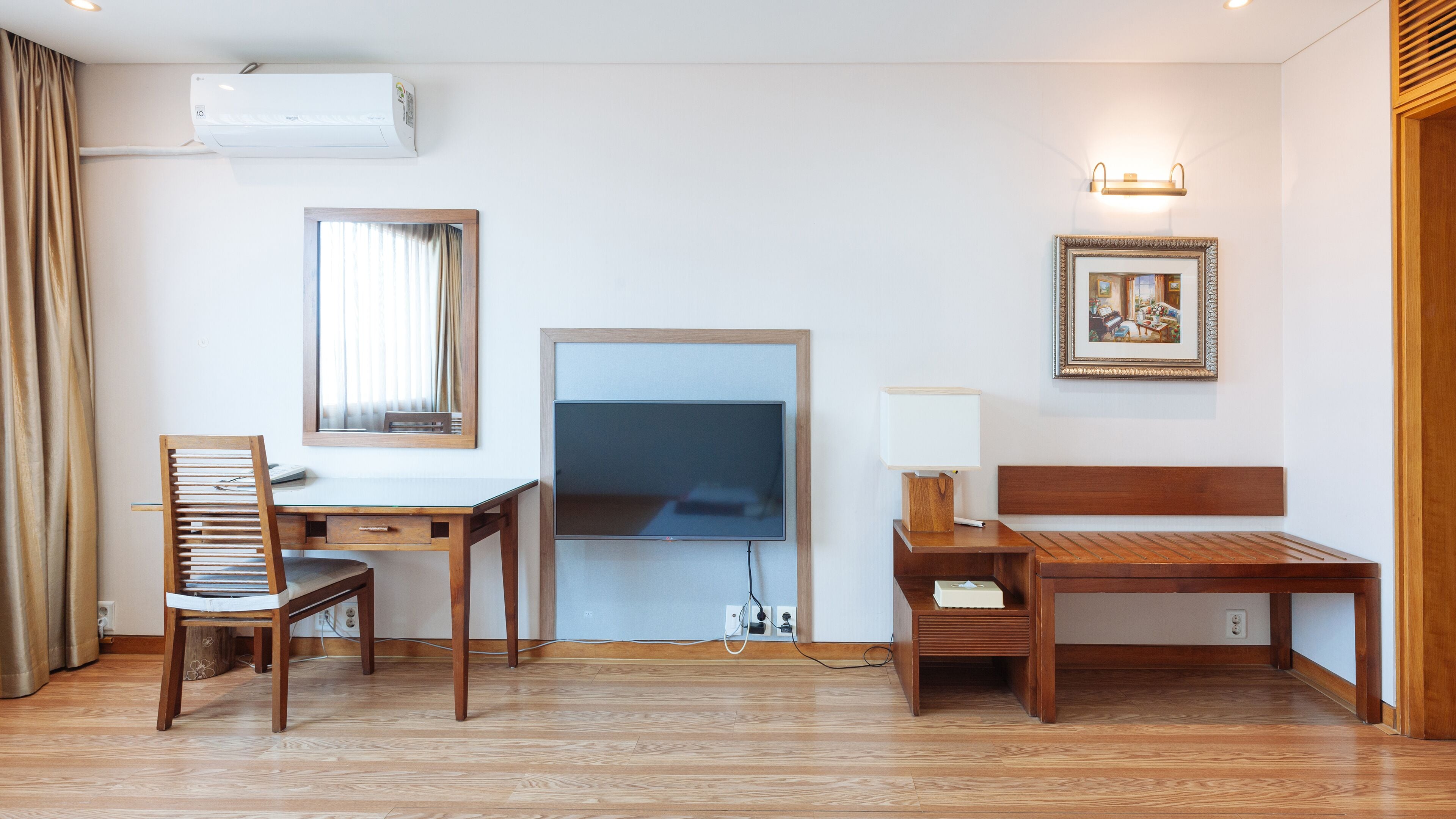 Habitación de hotel con un escritorio de madera, una silla, un televisor, una mesita de noche con una lámpara y un cuadro en la pared.