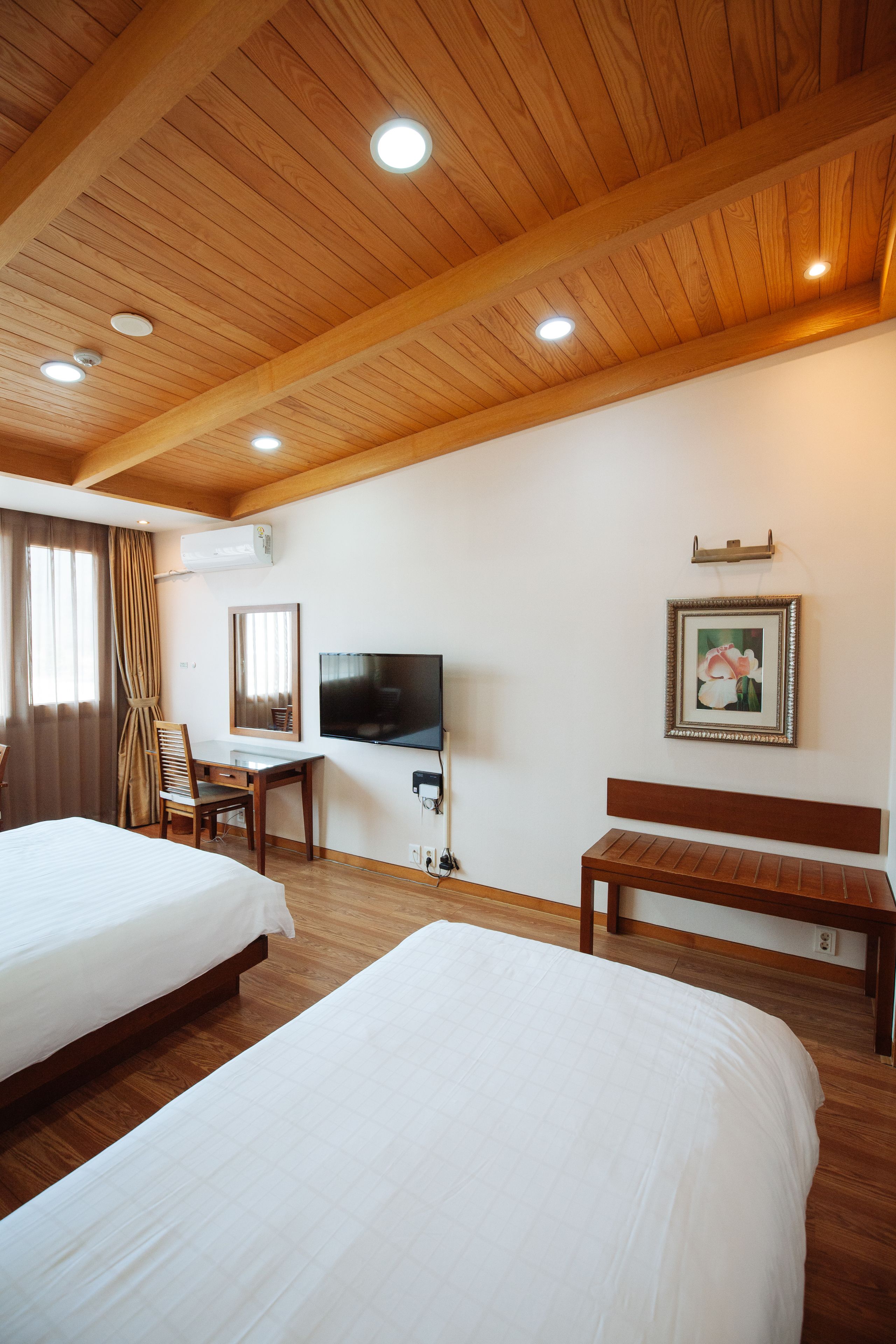 A hotel room with wooden ceilings, two beds, a desk, a TV, and a framed picture on the wall.