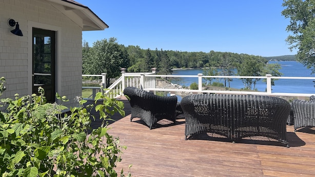A wooden deck with wicker furniture, a white railing, and a view of a lake surrounded by trees. - BEAUTIFUL OCEANFRONT COTTAGE WITH AMAZING WATER VIEWS & ACCESS (Hancock)