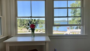 A room with a white table and bench, a vase with flowers, and a view of a lake and trees through two windows. - BEAUTIFUL OCEANFRONT COTTAGE WITH AMAZING WATER VIEWS & ACCESS (Hancock)