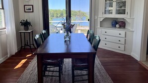 A dining area with a wooden table and chairs, a vase with flowers, and a view of a lake through a glass door. - BEAUTIFUL OCEANFRONT COTTAGE WITH AMAZING WATER VIEWS & ACCESS (Hancock)
