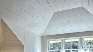 A bedroom with a sloped ceiling, a wooden dresser, a bed with a beige bedspread and a pink throw blanket, and a large window with a view of trees. - BEAUTIFUL OCEANFRONT COTTAGE WITH AMAZING WATER VIEWS & ACCESS (Hancock)