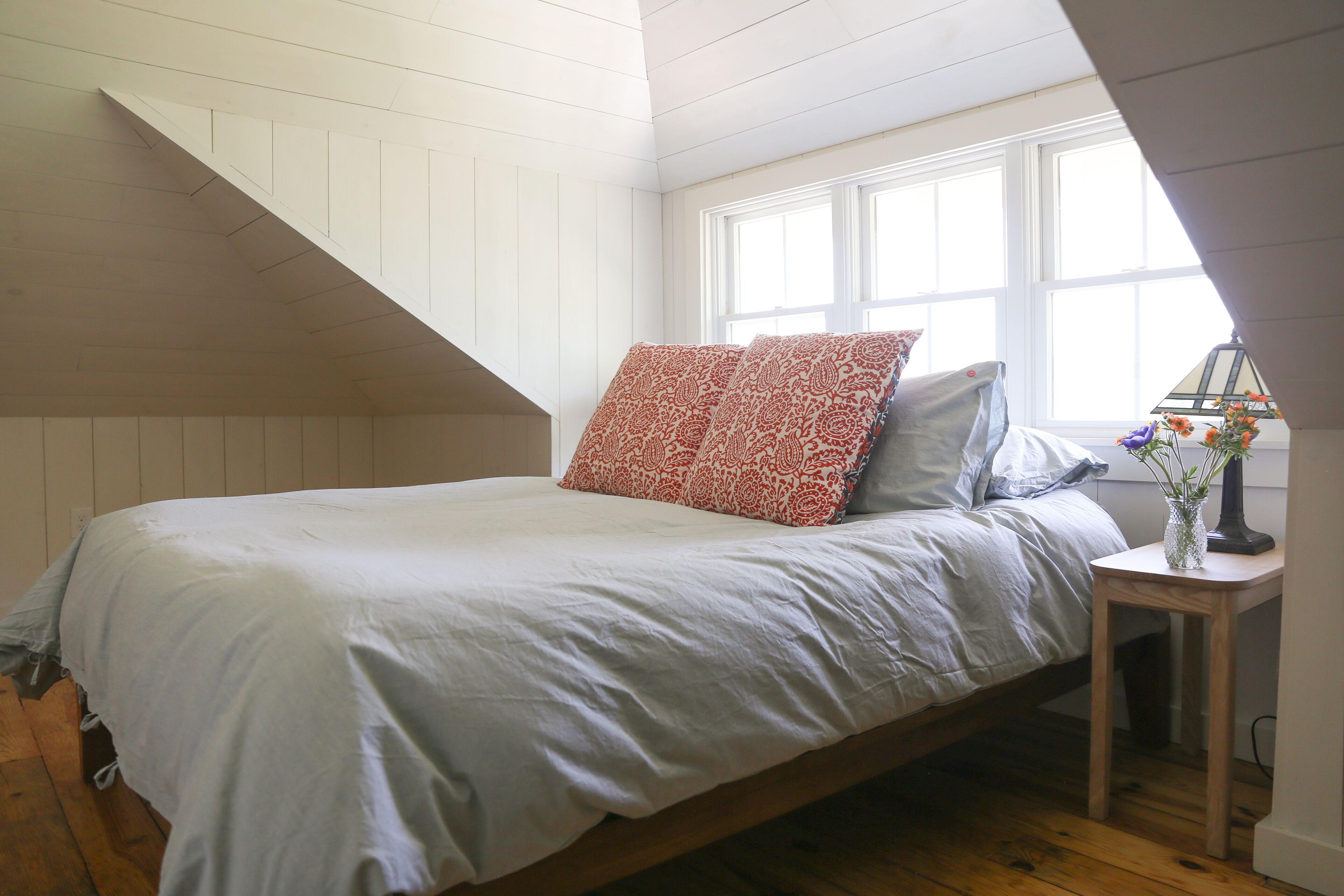 A bedroom with a sloped ceiling, a bed with patterned pillows, a nightstand with a vase, and a window with curtains.