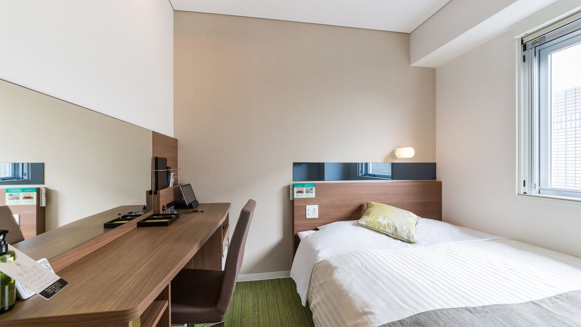 A hotel room with a bed, a desk with a chair, a mirror, a window, and a wall-mounted light.