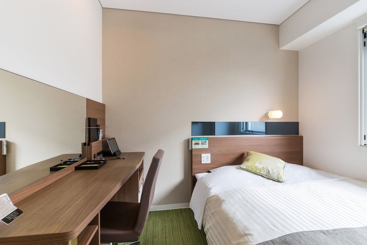 Ein Hotelzimmer mit einem Bett, einem Schreibtisch mit Stuhl, einem Spiegel, einem Fenster und einer Wandleuchte.