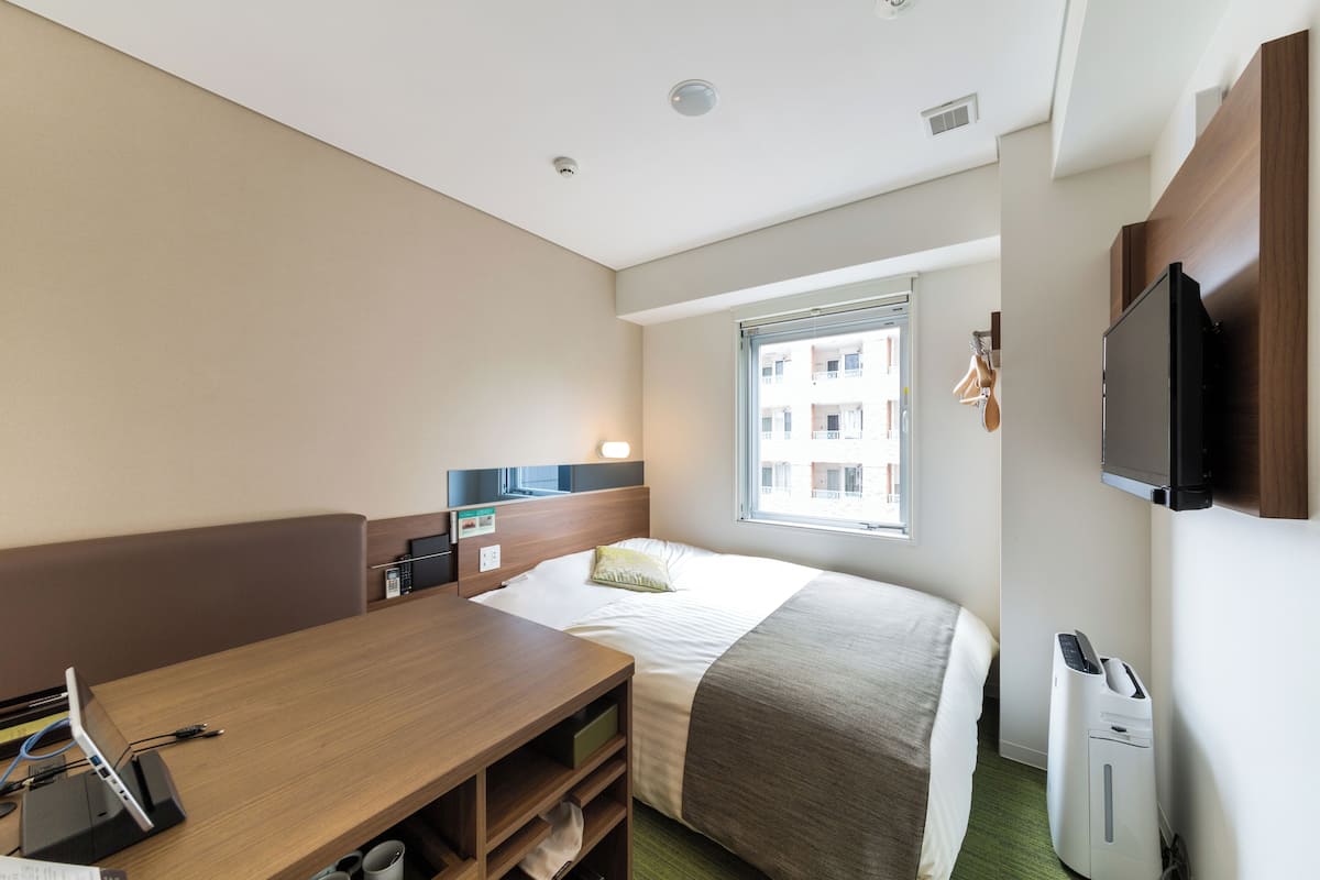 Ein Hotelzimmer mit Bett, Schreibtisch, Stuhl, Fernseher, Fenster und Mülleimer.