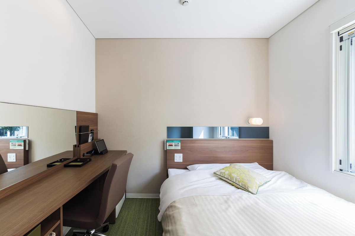 Ein Hotelzimmer mit einem Holztisch, einem Stuhl, einem Bett mit Kissen, einem Nachttisch, einem Spiegel und einem Fenster.
