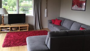 Un salon avec un canapé gris, des coussins rouges, un tapis rouge, un téléviseur posé sur un meuble en bois, une croix accrochée au mur et une fenêtre avec des rideaux.