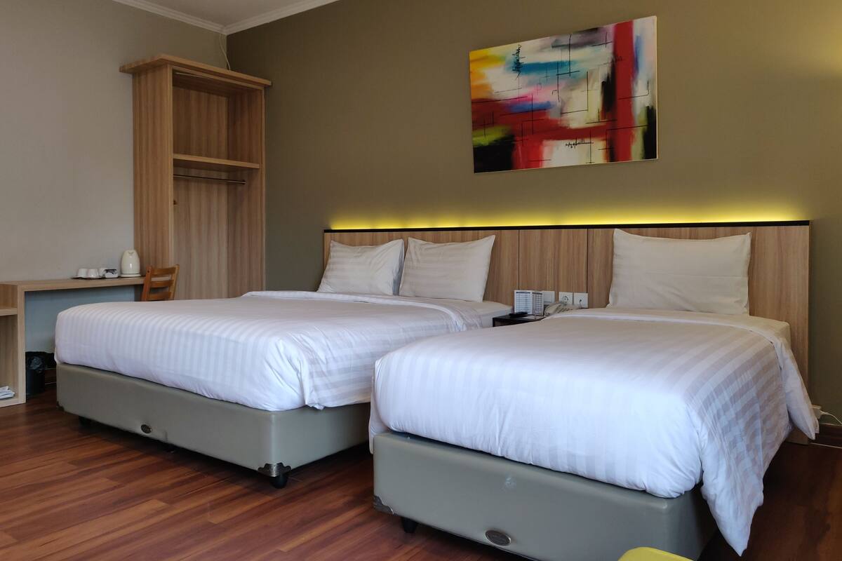 A hotel room with two beds, a wooden wardrobe, a desk with a chair, and a colorful abstract painting on the wall.